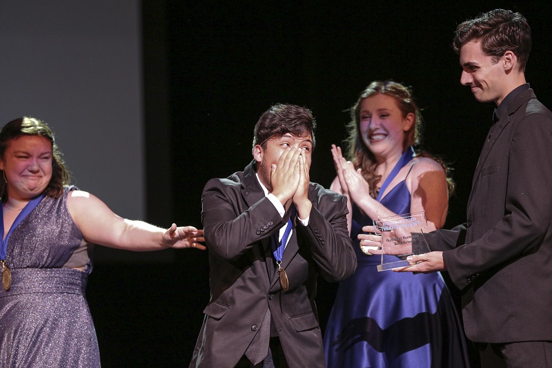 A young man in a suit is surprised on stage, while a group of his peers celebrate him.