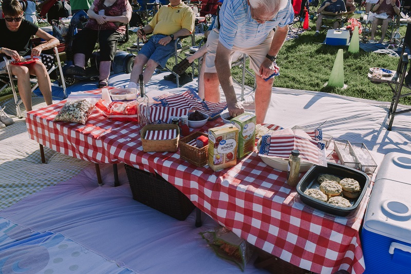 People grab food from a table adorned with a red and white checkered table cloth