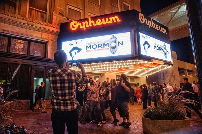 A group of people stand under the lit up Orpheum marquee before Book of Mormon. A person in the foreground takes a photo of them with a cellphone. Everything is smiling and excited.
