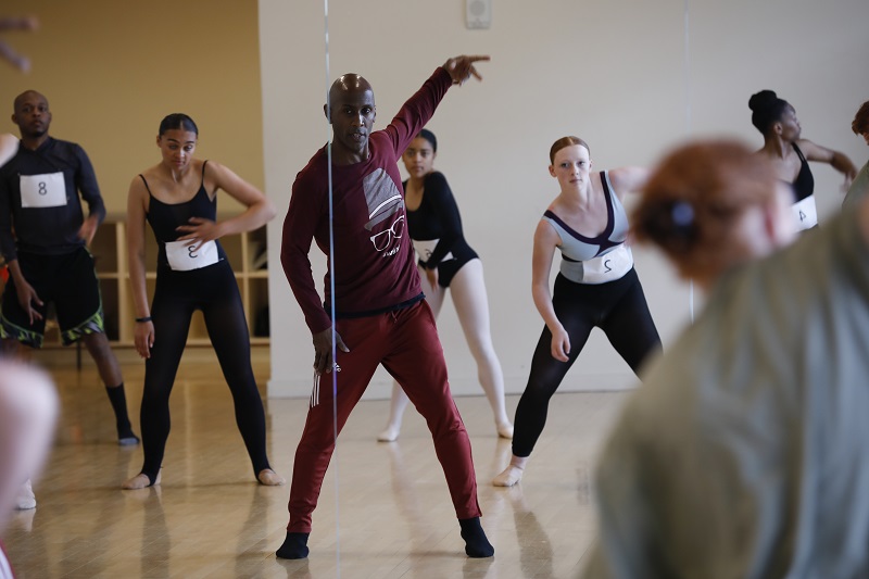 Ray Mercer stands in front of a mirror in a dance studio as dancers in leotards mimick his movements behind him