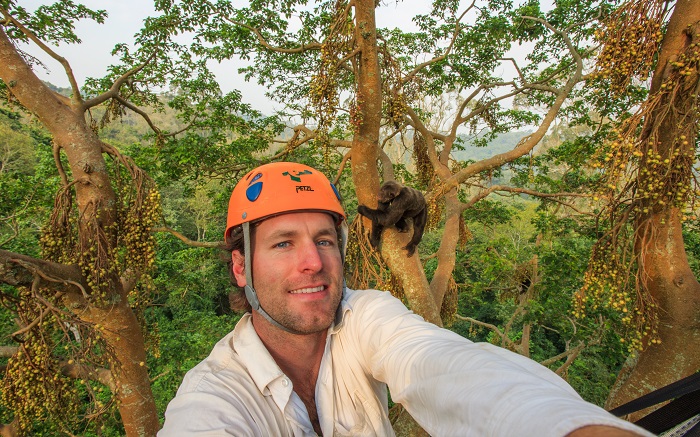 Ronan Donovan snaps a selfie of himself among jungle tree tops. In the background behind him, there's a chimp sitting in the street. Donovan wears an orange helmet. 