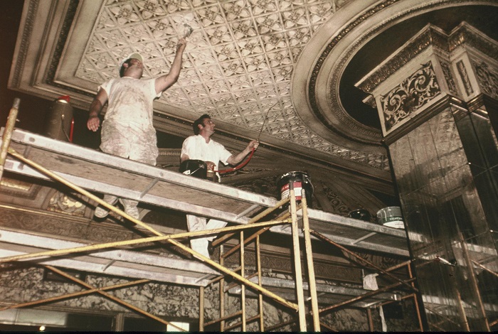 Two men stand on scaffolding to paint the faded ceiling of the Orpheum Lobby. The image is from 1974 and is faded. 