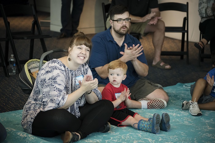 WeBop instructor and mom Kim Lomax sits cross-legged next to her toddler son. They're both clapping along to WeBop music with big smiles on their faces.